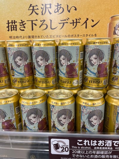 The image shows a close-up of several cans of "Premium YEBISU" beer with a unique manga-style design. The cans feature a female character illustrated by manga artist Ai Yazawa. They are arranged in rows. The cans are gold with a light blue design showing a woman in a kimono and flowers. A sign next to the cans reads "This is alcohol. 20 and older. You must confirm the age to buy alcohol". This photo was taken in Nakano, Japan.