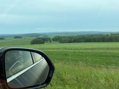 This media file captures a view from inside a moving vehicle, as seen through the side mirror and the window. The primary subject in the foreground is the side mirror of a black car, reflecting a person holding a smartphone and a portion of the car's interior. The smartphone has an orange case with stickers. The background depicts a vast rural landscape under an overcast sky, suggesting a cloudy or possibly rainy day. Rolling green fields stretch into the distance, dotted with clusters of trees and a faint treeline on the horizon. The sky is a muted gray-blue with some faint linear cloud formations or contrails. The image appears to have been taken during daylight hours. The overall mood is one of travel through a serene, green countryside. There is no visible text in the image. The location context provided is Mingul, Russia.