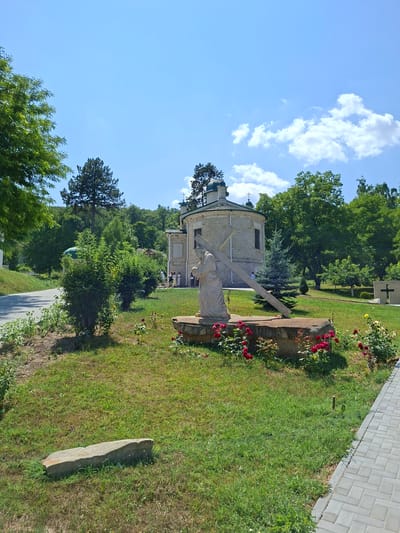 The image depicts an outdoor scene with a religious theme at the Condrița Monastery in Moldova. In the foreground, a life-size stone sculpture of Jesus carrying the cross is positioned on a stone base, surrounded by red roses. To the right, a paved walkway runs along a grassy area. In the mid-ground, a light-colored, rounded church with a green dome is visible. Several individuals, some wearing religious attire, are standing near the church entrance. A large tree stands behind the church, and a dense line of trees forms the background. The sky is blue with scattered white clouds.