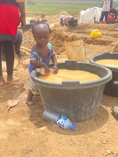 A photograph depicts a child filling a bottle with liquid from a large black tub in what appears to be an outdoor setting, likely in Durumi, Nigeria. The child, wearing a blue and green top and light-colored pants, is standing beside the tub, which is filled with a beige-colored liquid. A second, similar tub is partially visible behind the first. A bottle of water rests on the ground next to the tub. Other individuals are partially visible in the image, with legs and feet visible in the foreground. The ground is dry and dusty. In the background, there's a landscape with green fields.