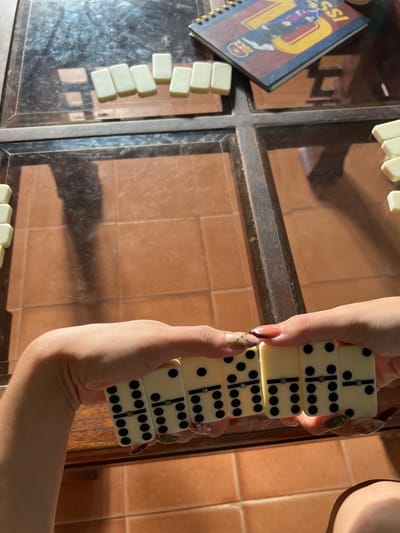 In Villa de Cura, Venezuela, an overhead, close-up shot captures a game of dominoes in progress on a glass-topped table. The table's surface reflects the terracotta tiled floor below and a shadow of a person. Several dominoes are scattered on the table, some in a row near the top left and others on the right side. In the foreground, two hands are holding a sequence of five dominoes, each displaying dots representing numbers. The dominoes are off-white with black dots. The hands show manicured fingernails with intricate designs. A spiral-bound notebook with a design featuring soccer player Lionel Messi is visible on the upper right side of the table.
