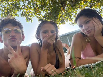 Three young people are lying on grass under a tree on a sunny day. A young man on the left is wearing glasses and making a peace sign with his left hand. A young woman in the center is covering her mouth with her right hand. A young woman on the right is resting her head on her hand and wearing a pink bikini top. They appear to be at a park or outdoor recreational area in Villa del Prado, Spain.