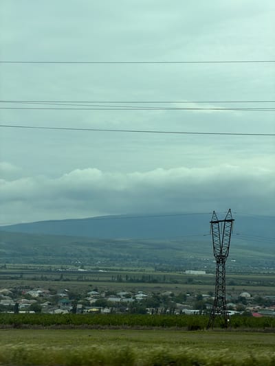 The image shows a landscape with power lines and a transmission tower. In the background, there is a valley or plain with a settlement or village. The scene is characterized by a combination of natural and industrial elements. The sky is overcast. The location is Tserovani, Georgia.