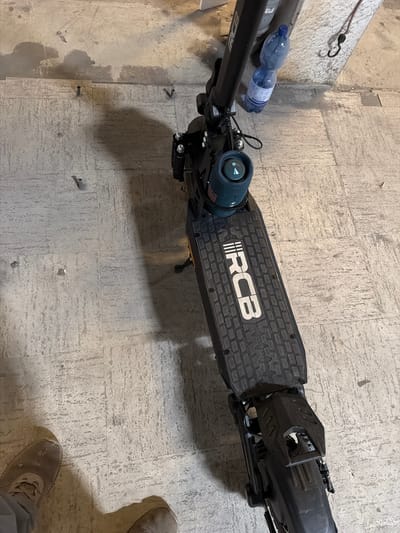An electric scooter is parked on a concrete floor with grey tiles. A blue water bottle is attached to the scooter's handlebars. The scooter has the "RCB" logo on its deck. A person's feet wearing casual shoes are visible in the bottom left corner of the image. The scene appears to be indoors, possibly in a garage or storage area. There is no identifiable location in the city of Osnabrück, Germany within the image.