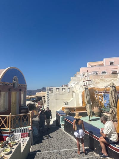 The image depicts a sunny day in Oia, a municipal unit of Santorini, Greece. The scene captures a narrow cobblestone street lined with traditional white-washed buildings, characteristic of the Cycladic architecture. The buildings feature colorful accents, including a prominent blue arched roof on the left and pink structures on the right.
In the foreground, to the left, a shop displays various ceramics and souvenirs on a low wall. Further up the street, several people are walking. A couple, a man and a woman, are seen walking away from the camera. The woman wears a grey dress and sunglasses, while the man wears a black t-shirt and shorts.
To the right, a woman wearing a straw hat and white shorts is walking towards the left, facing away from the camera. A man is seated on a low wall on the right, wearing a white polo shirt and blue shorts. He appears to be looking at something in his hands.
Several closed umbrellas are visible, suggesting a cafe or seating area. In the background, more buildings ascend the hillside, with glimpses of the Aegean Sea and distant islands under a clear blue sky. The overall impression is one of a bustling tourist destination in a picturesque Mediterranean setting.