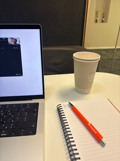 An overhead view depicts a workstation on a white table in an indoor environment, specifically identified as Nes Ziona, Israel. A silver Apple MacBook laptop occupies the left side of the table, displaying a video stream on its screen featuring a man with headphones, partially identified as "Aviad Hershkov." The laptop's keyboard prominently displays Hebrew characters. To the right of the laptop, a light beige disposable coffee cup, bearing the text "Espresso Presto Coffee" and "נס ציונה" (Nes Ziona) in Hebrew, stands upright. Minor liquid residue is visible on the cup's inner rim. Further to the right, a spiral-bound notebook with white ruled pages and a red spine is open, with handwritten text partially legible as "Job Exp" and "Company." An orange pen, marked with a white "Moovit" logo, rests diagonally across the notebook. The background consists of an out-of-focus dark gray upholstered surface, likely a wall or furniture, with a reflection of a light-colored wall and a vertical frame visible on the far right.