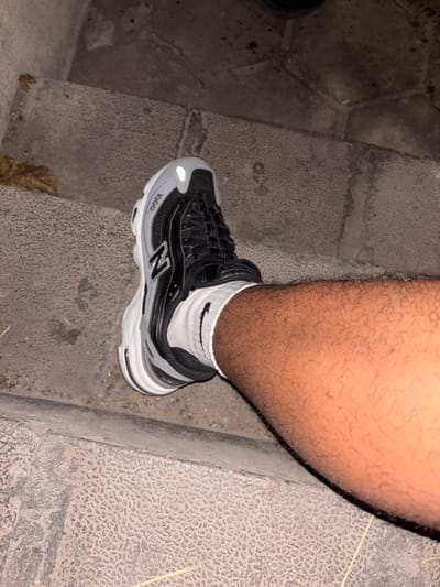 A close-up, eye-level shot captures a New Balance 1000 series sneaker worn on a person's right foot, positioned on textured concrete steps, possibly in Dimovo, Bulgaria. The shoe features a predominantly black and grey color scheme with white accents and a prominent "N" logo on the side. The laces are black, and the sock is white, pulled up to just below the ankle. The wearer's leg, covered in dark hair, is visible next to the shoe. The steps are made of grey concrete with a distinctive raised pattern and show signs of wear, with some debris scattered on them. The lighting suggests it may be evening or a dimly lit area.