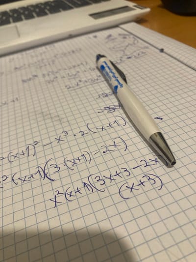 A white pen is laying on a sheet of graph paper with mathematical equations written in blue ink. The paper is sitting on a wooden surface. The location is Kokhav Ya'ir Tzur Yigal, Israel at coordinates 32.219, 34.991. The pen is laying horizontally on the paper near the bottom right corner. There are no visible people in the scene. The environment is a typical study or work space. The equations are handwritten in blue ink on the graph paper. The pen is a basic white pen. The surface of the paper is smooth.