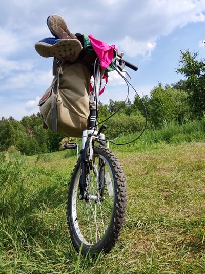 The image shows a close-up of the front of a bicycle parked in a grassy field under a cloudy blue sky. The bicycle has a tan canvas bag hanging from the handlebars, and a pink garment is draped over the bag. Two pairs of athletic shoes are resting on top of the bag, with their soles facing outward. The foreground is dominated by tall green grass, with a dense line of trees visible in the background. The lighting suggests it is daytime, and the scene appears to be in a rural or park-like setting. There is no indication of the city of Snopok Novyi, Russia in the image.