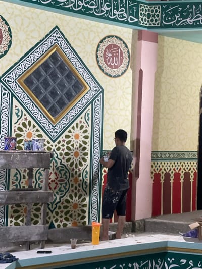 An image shows a man on a partially constructed scaffold working on a wall. The wall is decorated with Islamic art and includes a window with multiple square panes. The man is wearing a dark t-shirt and dark shorts with paint splatter on them. The wall is light yellow and green and the decor features patterns.