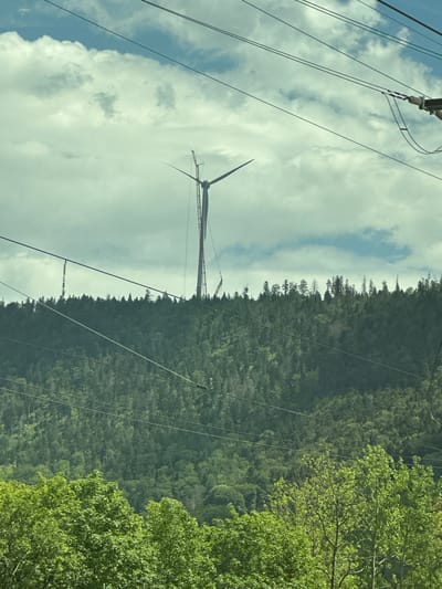 The image depicts a partially constructed or damaged wind turbine atop a densely forested hillside under a cloudy sky. The turbine's tower appears to be red and white, with sections of its structure exposed. One of the turbine blades is visible and seems to be leaning precariously, suggesting a potential structural issue or ongoing construction work. A crane is positioned near the top of the turbine, likely involved in its assembly or repair. Several power lines crisscross the foreground and extend across the image, obscuring parts of the sky and the hillside. The lower portion of the image features lush green trees, indicating a natural environment. The scene is devoid of people. The location is identified as Le Day, Switzerland.