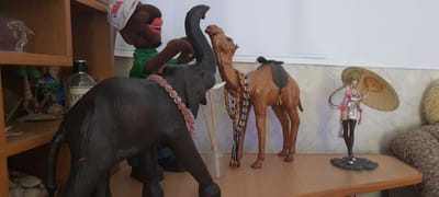 The image displays a collection of decorative items on a wooden shelf. In the foreground, a large, dark-colored elephant figurine with a pink beaded necklace is positioned on the left. To its right, a tan camel figurine with beaded necklaces and a saddle is standing. A puppet with a green shirt and a white hat with the NBA logo sits atop the elephant. To the far right, a figurine of a young woman in a pink outfit holding a large umbrella stands on a grey base. Behind the figurines, a light-colored, patterned wallpaper is visible. On the shelf to the left, a clear bottle with a black cap and a label reading "EYE" is partially visible, along with some smaller decorative items. The overall scene appears to be an indoor display of collectibles.