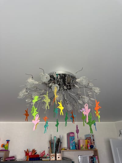 A close-up, eye-level shot shows a chandelier with multiple hanging glittered ballet dancer ornaments. The chandelier has a metallic base and decorative white flower-like accents. The dancers are in various poses and are colored pink, green, orange, yellow, and teal. The background consists of a white ceiling and a shelf unit with various items. The overall scene is indoors, likely in a room. The image does not contain any specific geographical markers to indicate its location in Мариинский, Russia.