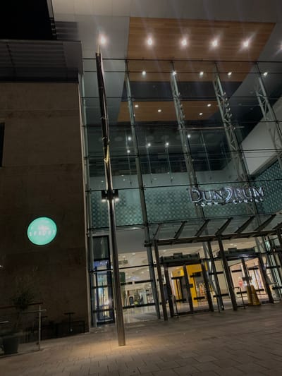 This is an exterior shot of Dundrum Town Centre in Dublin 16, Ireland, at night. The photo shows a multi-story structure with a glass facade displaying the "Dundrum Town Centre" sign. Below the glass facade is a stone-like wall with a circular, illuminated "Sprout" sign. The lighting appears dim, emphasizing the architectural lines and the signs. The exterior setting features a walkway paved with rectangular stone tiles. No people are visible in the scene.