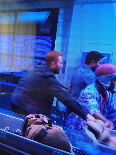 In Turka, Ukraine, a group of individuals is observed near a baggage claim area. A man with red hair is seen in the foreground, wearing a dark jacket and dark pants. To his right, a man with dark hair and a blue shirt is partially visible. Next to him, a person wearing a red head covering and a blue patterned garment is also present, with their hands near a light-colored bag. The environment appears to be an indoor public space, possibly an airport or train station, with bright blue lighting and visible textured surfaces on the walls and floors. The scene is captured at a slight upward angle, focusing on the people and the baggage carousel.