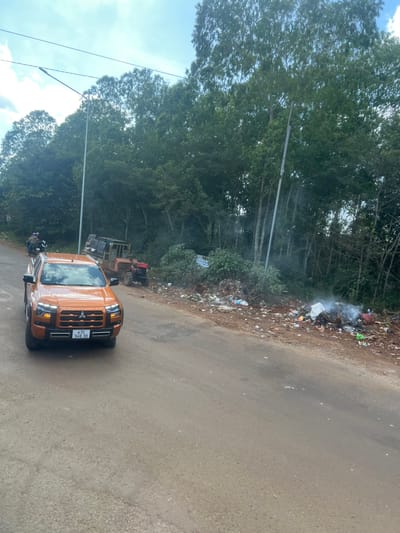 An orange truck occupies the lower-right corner of the frame, parked on an asphalt road. Green trees line the left side of the road and extend into the background. A pile of trash is visible along the roadside, emitting smoke. The scene appears to be on the outskirts of a forested area. A bulldozer is partially visible behind the truck.