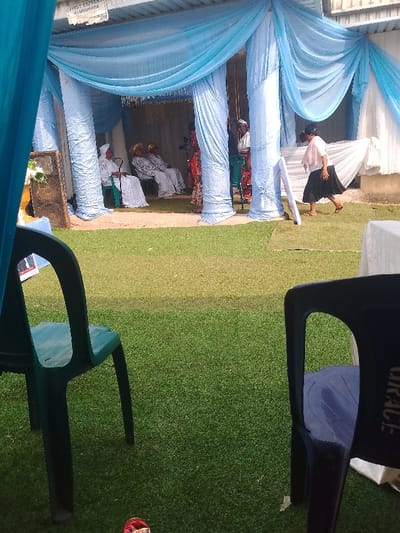 The image portrays an outdoor event setting in Ufuma, Nigeria. The ground is covered with artificial green turf. A temporary canopy structure dominates the background, featuring light blue fabric drapes and supporting columns also wrapped in light blue material. A metal sign, "FIRST EXPRESS ALUMINIUM," is visible at the top left of the structure.

Underneath the canopy, on the left side, three women are seated on plastic chairs. They are dressed in white traditional attire, including headwraps. To their left, a large, dark speaker with a small white floral arrangement is positioned. In the center-right section of the canopy, a woman in a red patterned dress stands, and another individual is partially visible behind a blue-draped column.

To the right of the sheltered area, a woman wearing a light-colored blouse and a dark skirt is captured mid-stride, walking from right to left across the artificial turf. She passes a white, upright sign board. A table draped with a white cloth is partially visible on the far right. In the foreground, a dark teal plastic chair occupies the lower left, and a dark blue plastic chair, marked with "GRACE" on its back, is present in the lower right. A single red sandal is visible at the bottom center of the frame.