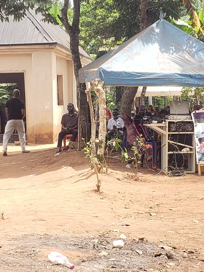 This image depicts an outdoor gathering in Umundugba, Nigeria, under bright daylight conditions. The scene is set on dusty, reddish-brown ground with ample green foliage and trees in the background.
On the right side of the frame, several individuals are seated on a mix of plastic chairs (orange and blue) beneath a blue tarp that serves as a makeshift shelter. Behind these seated people, a complex sound system or electronic setup is visible on a stand, featuring numerous wires and various components. A banner or poster, displaying what appears to be a portrait of a person, is affixed above the sound equipment.
In the mid-ground, a man dressed in a dark shirt and dark trousers sits on an orange chair, looking towards the viewer. To the left and slightly closer to the foreground, another man, wearing a dark shirt and light-colored trousers, stands with his back to the camera, near a light-colored building. This building has a dark window or doorway and a corrugated metal roof, partially obscured by tree branches.
The ground shows patches of varying texture and color, and a white plastic bottle with a pink cap is discernible among the debris in the foreground. Strong shadows cast by the sun indicate it is likely midday or early afternoon. The overall impression is of a community event or informal meeting in a rural or semi-urban environment. No explicit text is clearly legible in the image.