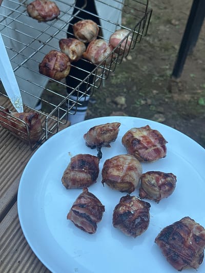 In Novaya Vozhoika, Russia, a close-up, high-angle shot captures a rustic outdoor setting where food is being prepared. A white plate, filled with approximately eight pieces of food wrapped in bacon, sits on a wooden surface. These bacon-wrapped items appear to be small, spherical food items, possibly meatballs or small potatoes, and are cooked to a golden brown with some charring. To the left of the plate, a metal grill basket is positioned, holding several more of the bacon-wrapped food items. A metal spatula is visible, its blade resting on the grill basket, suggesting an action of food preparation or serving. The background is out of focus, showing a natural, earthy environment with green vegetation and dark soil, consistent with an outdoor setting in Russia.