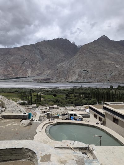 The image depicts an outdoor hot spring in Panamik, India, nestled amidst a rugged, mountainous landscape under a cloudy sky. In the foreground, a kidney-shaped pool filled with cloudy, greenish water is the central feature, surrounded by concrete and stone construction that appears to be in a state of partial completion or repair. Tools, including a wooden-handled object resembling a spade or hoe, are visible on the concrete.
Beyond the hot spring, a collection of small, traditional buildings with flat roofs are scattered across the terrain. A light-colored utility vehicle and a blue piece of equipment are parked near one of these structures. Further in the background, a wide, arid riverbed stretches out, bordered by lush green vegetation, including a line of tall trees. The middle ground transitions to a verdant valley with more trees and small clusters of buildings.
Dominating the background are immense, imposing mountains with jagged peaks, their rocky surfaces textured and eroded by time. The overall impression is one of a remote, natural setting enhanced by man-made structures. The cloudy sky casts a diffused light over the scene, contributing to a subdued color palette. No individuals are clearly identifiable as actively participating in any activity within the frame.