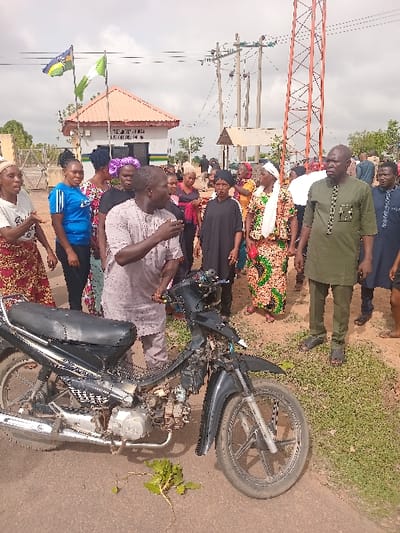 The image depicts a scene in Ungwan Tanko, Nigeria (latitude: 10.422, longitude: 7.49). A group of people, mostly women, are standing in a circle around a black motorcycle. They appear to be engaged in conversation. There is a small building behind them, possibly a police station or checkpoint. Two Nigerian flags are visible in the background, and a tall, orange communications tower is also visible. The ground is dirt, and there is a scattering of green vegetation. The atmosphere is calm and casual, with no indication of any conflict or tension.