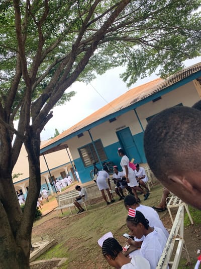 In Mkar, Nigeria, a group of people, predominantly women in white nurse uniforms with red and white striped caps, are gathered in the courtyard of a building with a corrugated iron roof and blue trim. The building appears to be a health facility or educational institution. The ground is covered with dirt and patches of moss. A large tree with dense foliage dominates the left side of the frame, casting shadows. Some individuals are seated on white benches, one woman is looking at her phone, while others are standing and walking. A bicycle is parked near the building. The overall scene suggests a break or gathering at an outdoor location.