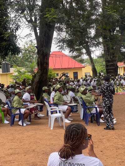 In Orun Ekiti, Nigeria, a drum major in a camouflage uniform directs a young band in green uniforms. The band members, seated on chairs, play drums and brass instruments. They are positioned outdoors under large trees, with a yellow building and a red-roofed structure in the background. A young woman in the foreground, seen from behind, is taking a photo with her phone. The ground is covered in red earth, suggesting a rural or semi-rural setting.