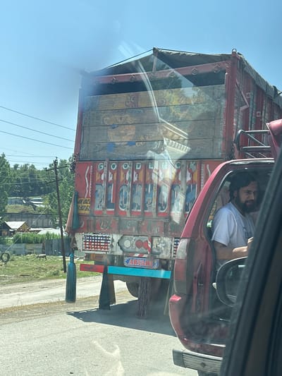 In Khrewan Lasi Pora, India, a red car is stopped on a paved road, its passenger-side door open. Inside the car, a man with a beard and wearing a white t-shirt is visible. Behind the car, a large, ornately decorated truck is parked. The truck's red and wooden body features colorful paintings and text. A teal-colored tasseled ornament hangs from the truck's rear. In the background, trees and buildings are visible under a clear blue sky. A power pole with wires stands to the left of the truck, and a few animals are in a grassy field behind a fence.