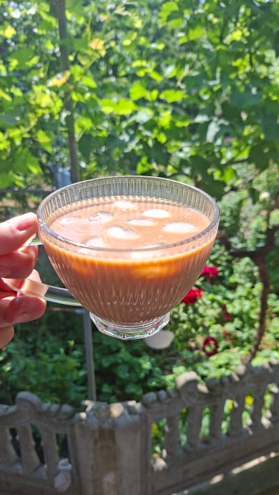 A hand holds a clear glass mug filled with a brown beverage and ice cubes. The mug has a ridged texture and a clear handle. The background is a blurred garden scene with lush green foliage and some red flowers. A decorative concrete fence is visible in the foreground. The image appears to be taken outdoors on a sunny day. The location is identified as Bălășești, Moldova.
