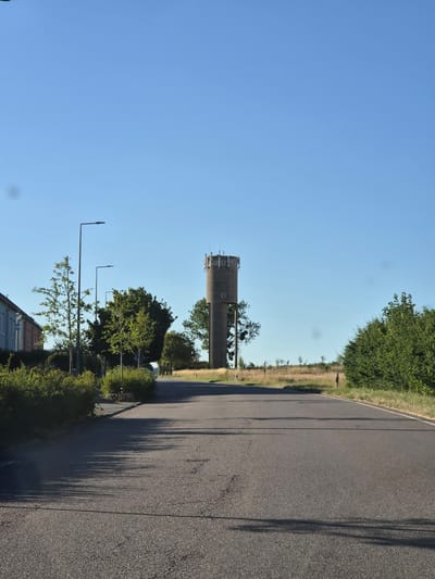 This is a vertical, eye-level, full shot of a rural scene with a water tower in the background. The sky is clear and blue, indicating fair weather. The sun appears to be setting, casting long shadows across the asphalt road that dominates the right side of the frame.
The main subjects are the water tower, a road, trees, and streetlights. The water tower is a tall, cylindrical concrete structure with visible windows and some antennae or equipment on top. It is situated in the distance, nestled amongst a line of trees and vegetation. The road is paved and appears to be a two-lane rural road with a white line marking the edge. Lush green trees and bushes line the left side of the road, with a field of dry grass and scattered trees further in the background. Several modern streetlights are visible along the side of the road, some with their lights illuminated. In the foreground, the roof of a building is partially visible on the lower left.
There are no people or animals visible in the image. The overall mood is peaceful and serene, characteristic of a quiet rural evening. The presence of the water tower and the road suggest a developed area within a rural landscape. The location is specified as Schengen, Luxembourg. No visible text is present in the image.