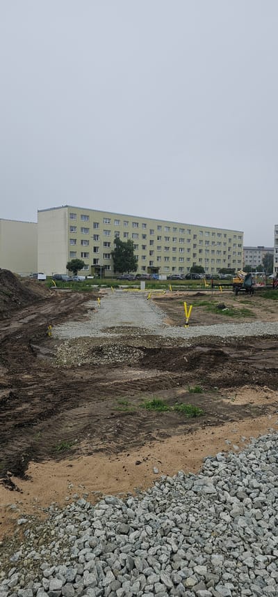 This is a photograph taken outdoors on a cloudy day, showing a construction site in front of a long, pale yellow, multi-story apartment building. The apartment building has many windows, arranged in a grid pattern. There are several cars parked in front of the building, and some trees.

The foreground of the image is a construction area with disturbed earth, tire tracks from heavy machinery, and piles of gravel. Yellow markers or pipes are visible sticking out of the ground in various locations, suggesting ongoing utility work or ground preparation. A small yellow excavator is parked in the middle ground, near the apartment building.

The apartment building appears to be in the city of Maardu, Estonia, and the overall scene suggests a renovation or new construction project is underway in a residential area. There are no people clearly visible in the photograph.