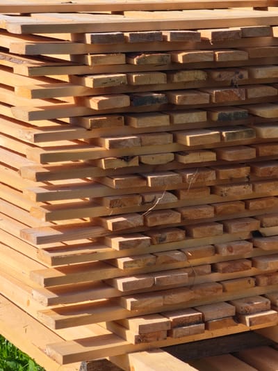 A stack of sawn lumber, predominantly pine, is piled in an outdoor setting. The wood planks are of varying thicknesses and lengths, with some showing rough-sawn edges and natural wood grain. The lumber is stacked horizontally in several rows, creating a staggered effect. The wood exhibits a light to medium brown color, with some pieces showing darker tones from sapwood or weathering. Sunlight casts distinct shadows across the planks, highlighting their texture and form. In the lower left corner, a patch of green grass is visible, suggesting the lumber is stored outdoors, possibly in a construction site or lumber yard. The image does not depict any people or animals. The overall impression is one of raw building materials, organized for storage or transport. The location in Zhukovo, Russia, is a detail provided outside the visual information of the image.