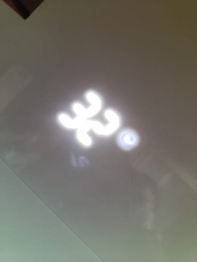 The image shows a light reflection on a smooth, light-colored surface, possibly a wall or ceiling. The reflection appears to be in the shape of a stylized symbol, with four curved arms radiating from a central point. There is also a smaller, circular light reflection to the right of the main symbol. The overall impression is of a bright light source casting a patterned reflection. The location in Zarubino, Russia is not discernible from the image.