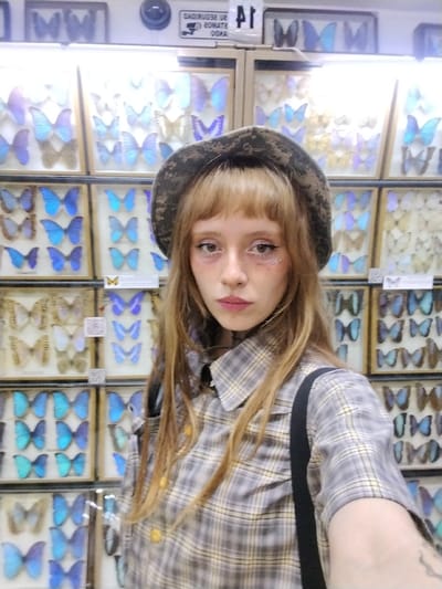 A young person, likely female, is featured in a close-up, slightly off-center selfie taken indoors. She has fair skin, light-colored eyes, and long, straight light brown hair with bangs that fall across her forehead. Her makeup includes winged black eyeliner, a reddish tone beneath her eyes, and pinkish-mauve lipstick. Small, shiny embellishments, possibly gems or glitter, are visible on her left cheekbone. She wears a neutral expression, looking directly at the camera.
She is dressed in a short-sleeved plaid button-up shirt featuring shades of grey, beige, and yellow, with yellow buttons down the front. A dark, possibly black, strap from a bag or garment is visible over her right shoulder. On her head, she wears a bucket hat with a camouflage or earthy abstract pattern in muted tones.
The background consists of multiple stacked display cases, likely part of an exhibition. These cases are filled with meticulously arranged and pinned butterflies. A significant number of the butterflies exhibit iridescent blue and purple wings, suggesting species like Morpho butterflies, while others are shades of brown, white, and lighter blues. The cases are illuminated by bright, even lighting.
Above the display cases, a white wall or ceiling is visible. A dark, circular security camera is mounted in the upper left of the background. To the right of the camera, a white sign with black text is partially visible. The text appears to read "CUIDADO SE" on the top line, and "OBSERVADO" with "20MA" or "2OMA" and "OSNA" or "GSMA" below it, strongly suggesting a warning sign in Spanish or Portuguese, likely "CUIDADO, SE ES OBSERVADO" (Caution, you are being observed). Another small, partially obscured sign next to it seems to display "P1" or "PL". Smaller white labels are present within the butterfly display cases, but their text is illegible.
The setting appears to be a museum or an entomological collection, showcasing preserved butterfly specimens. The person is posing for a photograph in front of these displays.