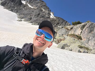 The image shows a man wearing sunglasses and a baseball cap, smiling, with a black jacket and backpack. The background consists of a mountain landscape with rocks, snow, and a clear blue sky. The location is the Rila Mountain, near the city of Govedartsi, Bulgaria. The photo is taken from an unusual angle, possibly upside down.