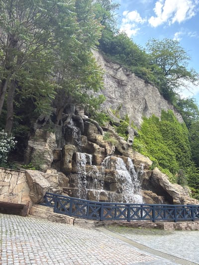The image shows a multi-tiered waterfall cascading down a rocky cliff face. The cliff is covered in lush green vegetation, including ivy. Trees with exposed roots are growing from the top of the cliff and overhang the waterfall. A dark blue wooden railing with an 'X' pattern runs in front of the waterfall, separating it from a cobblestone paved area. A wooden bench is visible to the left of the railing. The sky above is blue with scattered white clouds. This scene is located in Borjomi, Georgia.