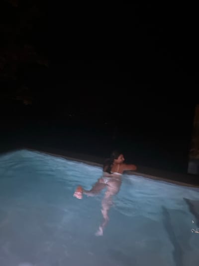 The image shows a woman in a white bikini swimming in a pool at night. The scene is located in Santa Fe, Costa Rica, at the coordinates 9.646, -85.166. The woman is in the water, with her head and upper body out of the water. She has long dark hair and is looking to the right side of the frame. The water is a light blue, reflecting the pool lights. The sky and surrounding area are dark. The pool edge is a light grey color.