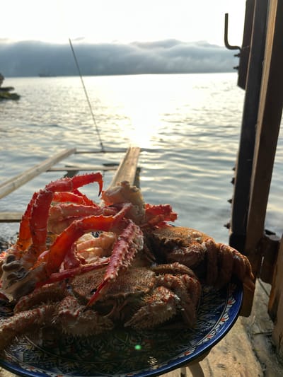 In Sovetskaya Gavan', Russia, at sunrise, a plate of cooked crab legs and whole crabs is presented on a wooden surface. The crabs, vibrant red and spiky, are arranged on a blue and white patterned plate. In the background, the calm sea reflects the warm glow of the rising sun, with a distant ship and a misty shoreline visible. A wooden structure and a fishing rod are positioned to the right and center of the frame respectively, adding to the outdoor, seaside ambiance. The scene suggests a fresh catch enjoyed at the water's edge.