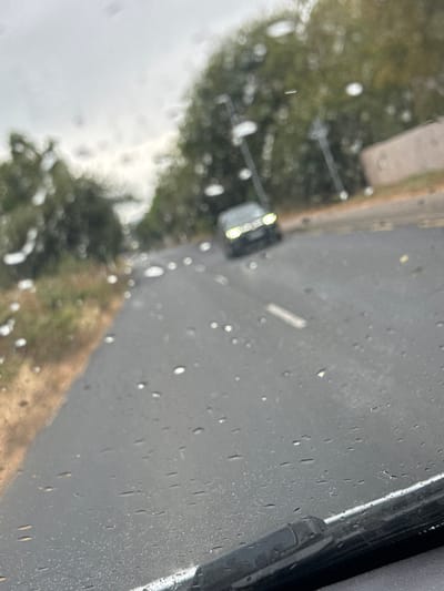 An overcast day in Mount Alexander, Ireland, as evidenced by the dull sky and the presence of raindrops on a car's windshield, which obstructs the view. The road ahead is wet and appears to be asphalt, with a dashed white line marking the center. In the distance, a dark-colored car with its headlights on is visible. The surrounding environment consists of a grassy verge on the left and trees and a building on the right, all blurred due to the rain and the perspective from inside the vehicle. The bottom of the image shows the edge of a windshield wiper.