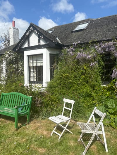 The image shows a white cottage with a green bench and two white folding chairs in front of it, set against a backdrop of lush greenery. The cottage is partially covered by a vine with purple flowers. There is a blue bottle on the green bench. The scene is in Balerno, United Kingdom, located at latitude 55.891 and longitude -3.326. The weather is sunny and the sky is blue with white clouds. The green grass is neatly manicured.  The windows are framed with black trim.  The roof is made of black slate tiles. There are two skylights in the roof. The chimney is red brick.  The front of the cottage is covered in white paint, with black accents.