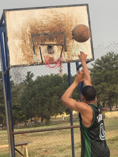 The image displays an outdoor basketball court in Umunekwu, Nigeria. A male individual, viewed from the back and right shoulder, is depicted in the act of shooting a basketball. He wears a black and green sleeveless jersey with "TATUM" and the number "0" visible in white on his back. Both of his arms are extended upwards towards the basketball hoop. A worn, brown basketball is airborne, positioned slightly above and to the right of the hoop's rim. The backboard is a rectangular, white-painted metal structure, extensively covered in brown rust, with the heaviest concentration around the rim's mounting area. The metal rim is rusted, and its attached net, composed of red and white fibers, is tattered and partially torn. The hoop is supported by blue metal posts. A chain-link fence separates the immediate playing area from the background. Beyond this fence, several small, indistinct human figures are visible in an open, grassy expanse. Numerous green trees form the backdrop, and a single bare tree branch is noticeable on the far left. A rudimentary wooden structure, possibly a bench or table, is partially visible in the lower-left foreground, behind the chain-link fence. The sky above is overcast.