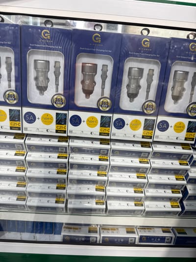 The image shows a retail display of mobile phone accessories, specifically car chargers and data cables. The products are neatly arranged in stacks, predominantly in their original packaging. The packaging is primarily dark blue with gold and white accents, featuring branding that includes a prominent "G" logo and the text "G-POWER" and "Dual Output Fast Charging Car Set". Some packages also highlight "365 DAYS WARRANTY" and specifications like "5V 5A DUAL USB" and "Type-C Data Line". The model numbers "C21-C" and "A66-C" are also visible. The background suggests a store or warehouse setting with shelves and fluorescent lighting, typical of a point-of-sale environment. There are no people visible in the image, and the overall impression is one of commercial organization and product presentation. The visible text on the packaging indicates the product type, brand, warranty information, and technical specifications. The location context of Bireuenmeunasah, Indonesia, is provided as external information.