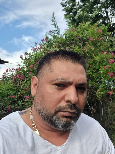 A close-up shot shows a man with short dark hair, a salt-and-pepper beard, and light skin. He is wearing a white t-shirt and a gold chain around his neck. The background includes green foliage with small pink flowers, blue sky, and white clouds. The image was taken in Zavoy, Bulgaria.