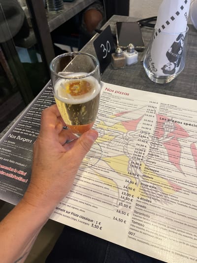 A restaurant table setting, presumably in Saint-Just-Saint-Rambert, France, features a partial view of a menu displaying a list of pizzas, including "Nos pizzas spéciales." A wine glass filled with a golden liquid, possibly wine or beer, is held by a hand with a visible tattoo. Additional table items include a salt and pepper set, a table number (30), and a wrapped object in a glass with a black and white image. The restaurant offers "Nos Burgers."