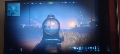 This is a screenshot from a video game, likely a first-person shooter, displayed on a television screen. The game appears to be set in a post-apocalyptic or war-torn environment, with a smoky, hazy atmosphere and buildings in disrepair. The scene is illuminated by a bright, fiery explosion in the background, suggesting an active conflict.
The player's perspective is through a sniper scope, with a reticle and crosshairs visible. A green laser sight is also projected, indicating the player is aiming at something specific. Below the scope, text in Russian reads "L3 - ГИБРИДНЫЙ РЕЖИМ," which translates to "L3 - HYBRID MODE." This suggests a game mechanic or mode being activated.
The user interface at the bottom of the screen displays game information. A rifle icon with the number "23" and "150" suggests ammunition count or weapon stats. Other icons and buttons (L1, R1) indicate controls. A mini-map in the top left corner shows a yellow marker, likely the player's current location or an objective.
The time of day appears to be dusk or dawn, with the sky exhibiting a blueish hue and the explosion providing a strong light source. The setting includes elements like power lines and what look like industrial structures. The overall mood is tense and action-oriented, typical of a combat simulation game. While Svitlovodsk, Ukraine is provided as a location context, there are no specific visual cues within the image itself to confirm this particular location.