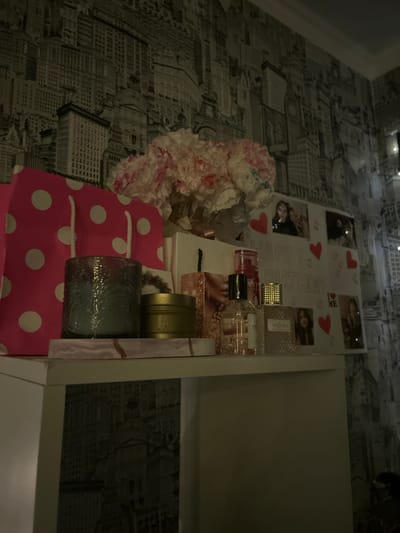 An indoor scene in Raduga, Russia, displays a collection of decorative and gift items arranged on a white shelf against a wall covered in black and white wallpaper depicting various cityscapes and famous landmarks. The lighting is dim, casting soft illumination on the objects.

From left to right on the shelf:
A large, fuchsia pink gift bag with white polka dots and white rope handles stands upright. To its right is a cylindrical, dark gray-tinted glass candle holder with an embossed pattern. Next to the candle holder is a small, metallic gold circular tin. Several perfume bottles are present, including a clear glass bottle with a black cap and gold collar, and another textured glass bottle with a gold cap and a label possibly reading "PANDORA." Behind these bottles, a cluster of light pink and white artificial flowers is visible.

A flat, white display board or card with the words "HAPPY BIRTHDAY" printed or written on it is positioned to the right of the flowers and perfume bottles. This display is adorned with several red heart cutouts and multiple small photographs affixed to it. These photographs depict individuals, primarily young females.

The environment suggests a personal space, possibly a bedroom, characterized by the celebratory arrangement and the decorative wallpaper. No active individuals are present in the scene.