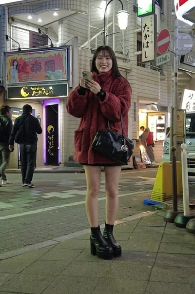 This nighttime image depicts an urban street scene in Gifu, Japan, specifically within the Miyasajimacho district, characterized by dense commercial buildings and entertainment venues.

In the foreground, a young East Asian woman stands centered on a paved sidewalk, smiling directly at the camera. She has long, dark hair with bangs and is wearing an oversized, textured dark red turtleneck sweater-coat, a black mini-skirt, and black platform boots with chunky heels. She holds a light-colored smartphone with a clear case in both hands at chest level. A black patent leather shoulder bag with a circular silver embellishment hangs from her left shoulder.

Behind her, on the left side of the frame, two men walk away from the camera. The man closer to the foreground wears a black jacket and jeans. Further back, another person in a reddish top is partially visible inside a brightly lit storefront on the right.

The background consists of multi-story buildings adorned with numerous illuminated signs. Prominently on the left, a large rectangular sign with a red background and gold border displays Japanese text ("必卓團"), an image of a woman, and a white animal. Beneath it, a black horizontal sign features a yellow crescent moon logo and Japanese text ("ななちゃん"). The entrance below this sign is illuminated with purple light.

To the right of the main subject, a vertical white sign with Japanese text ("たかはし") is visible, above which is a green rectangular sign. Below "たかはし," a smaller white sign shows "宮佐島町" (Miyasajimacho). Utility poles with overhead wires and streetlights illuminate the scene. On the right, a yellow traffic cone with a blue base stands on the sidewalk, and a portion of a white vehicle is visible. The street surface has visible road markings.