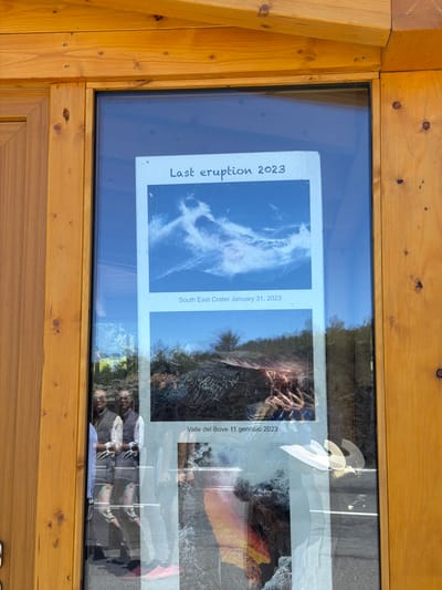 The image shows a glass-enclosed display on a wooden structure. Inside the display are two printed photographs. The top photograph depicts a sky filled with dramatic, white clouds against a blue background, with text below that reads "Last eruption 2023 South East Crater January 31, 2023". The bottom photograph shows a volcanic landscape with glowing molten rock and solidified lava, with text below that reads "Valle del Bove 11 gennaio 2023". In the reflection on the glass, two individuals are visible, wearing vests and shirts, and the photographer's hands are also reflected. The wooden structure appears to be part of a building or information kiosk, potentially in an outdoor setting given the reflections. The presence of text in Italian and the mention of "Valle del Bove" suggest the location is likely near Mount Etna, a volcano in Sicily, Italy, known for its eruptions and the Valle del Bove (Bove Valley). Linguaglossa is a town on the northern slopes of Mount Etna, making it a plausible location for such a display.