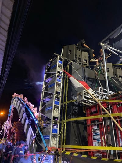 The image depicts an outdoor nighttime scene in Kedungbanteng, Indonesia, centered on a large, multi-tiered sound system. The structure is composed of numerous grey and black speaker enclosures, including several vertically stacked units branded "NEOERA" with blue illuminated panels. Two individuals are situated on the upper part of the sound system; one is seated with their back to the camera on a black speaker cabinet, while another is seated further right, facing left.

An Indonesian flag (red and white) is attached to the right side of the main sound system structure. Yellow safety straps are visible securing various components. To the right, a red-colored framework, possibly a vehicle or part of the support structure, features a sign with "TRUCK MANIA" and "PROTAPER" text.

On the left, an elaborate, arching decorative structure, possibly a parade float or art installation, extends upwards. It is adorned with white and light-colored elements and exhibits blue internal illumination, appearing to resemble a stylized animal or mythological creature. A crowd of multiple individuals is visible at the bottom left, gathered in front of the decorative structure and partially obscured by the foreground elements. Overhead, the sky is dark, indicating night. A street light is visible in the distant left background. A faint light beam or smoke effect emanates from the sound system towards the upper left.