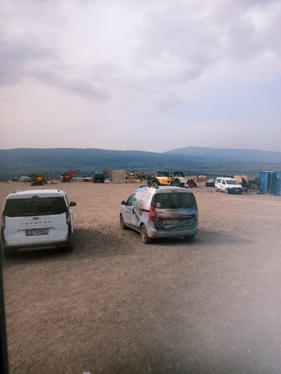 The image displays an open, unpaved industrial or storage yard situated in Drugan, Bulgaria, under an overcast sky with mountains forming the distant background. In the foreground, two vans are parked. On the left is a white Ford Tourneo Connect van, license plate CB 7654 EO, positioned with its rear facing the viewer. To the right, a light-colored van, resembling a Dacia Dokker, is parked at a slight angle, also with its rear facing the viewer. This van features a colorful geometric graphic wrap and carries license plate CB 7534 CL. The midground and background of the yard contain various pieces of heavy machinery and industrial materials, including an orange-red excavator, a blue utility truck, wooden crates, at least two large yellow construction vehicles (likely telehandlers or loaders), multiple large spools of cable, and stacks of blue industrial barrels. A white van is partially visible further in the background. No individuals are present within the frame.